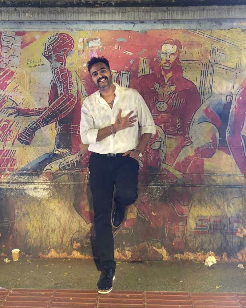 Amit in a white shirt, hand on his chest, laughing against a faded superhero mural on a Bangalore wall
