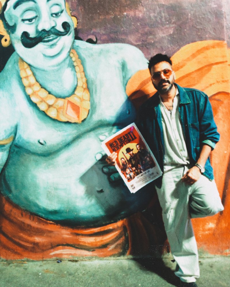 Amit leaning on a street mural of a painted giant with the same moustache, holding a poster