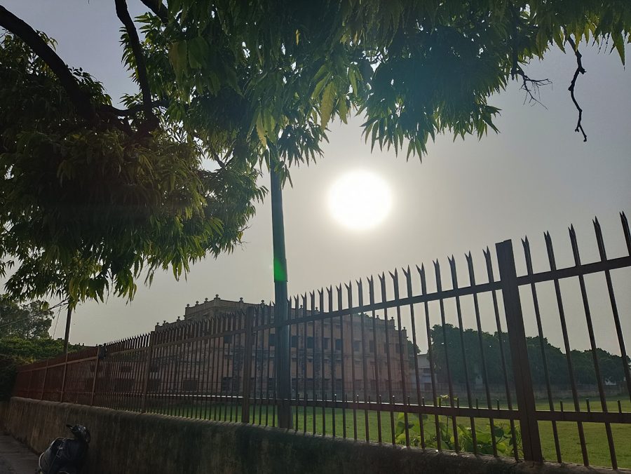 An old Firozabad building behind an iron fence, the sun in a hazy sky under neem leaves